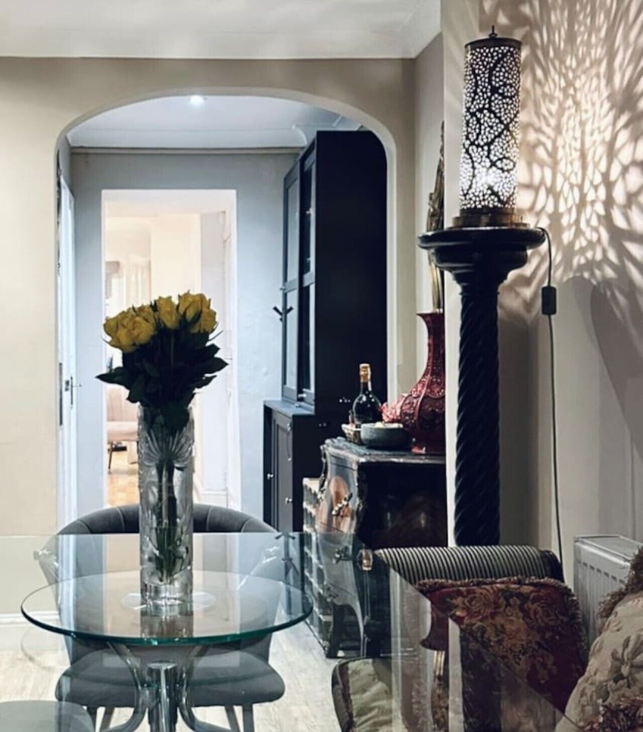 A dining area with a glass table, chairs, a vase with yellow flowers, a black cabinet, and a decorative wall piece.