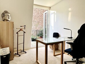 Une pièce avec un bureau en verre, une chaise, un porte-manteau suspendu, une statue de Bouddha et un balcon agrémenté de plantes.