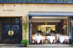 Un espace extérieur aménagé pour les repas, avec des tables dressées pour les clients, une grande porte décorée sur laquelle on peut lire le nom « DAPHNE'S », et un mur de briques en arrière-plan.