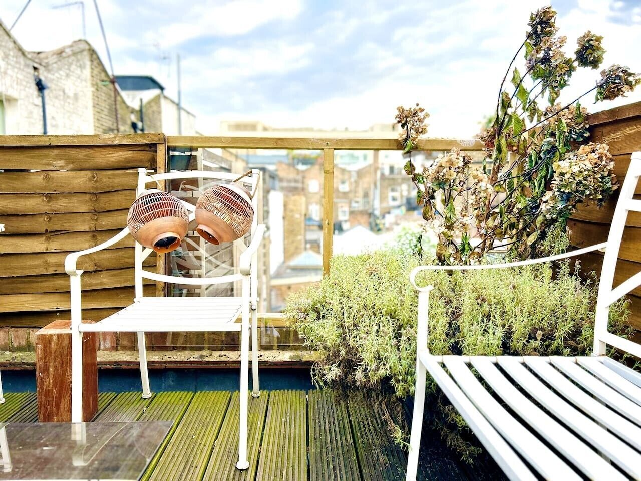 A wooden deck with two white chairs and a table, surrounded by plants and a brick wall.