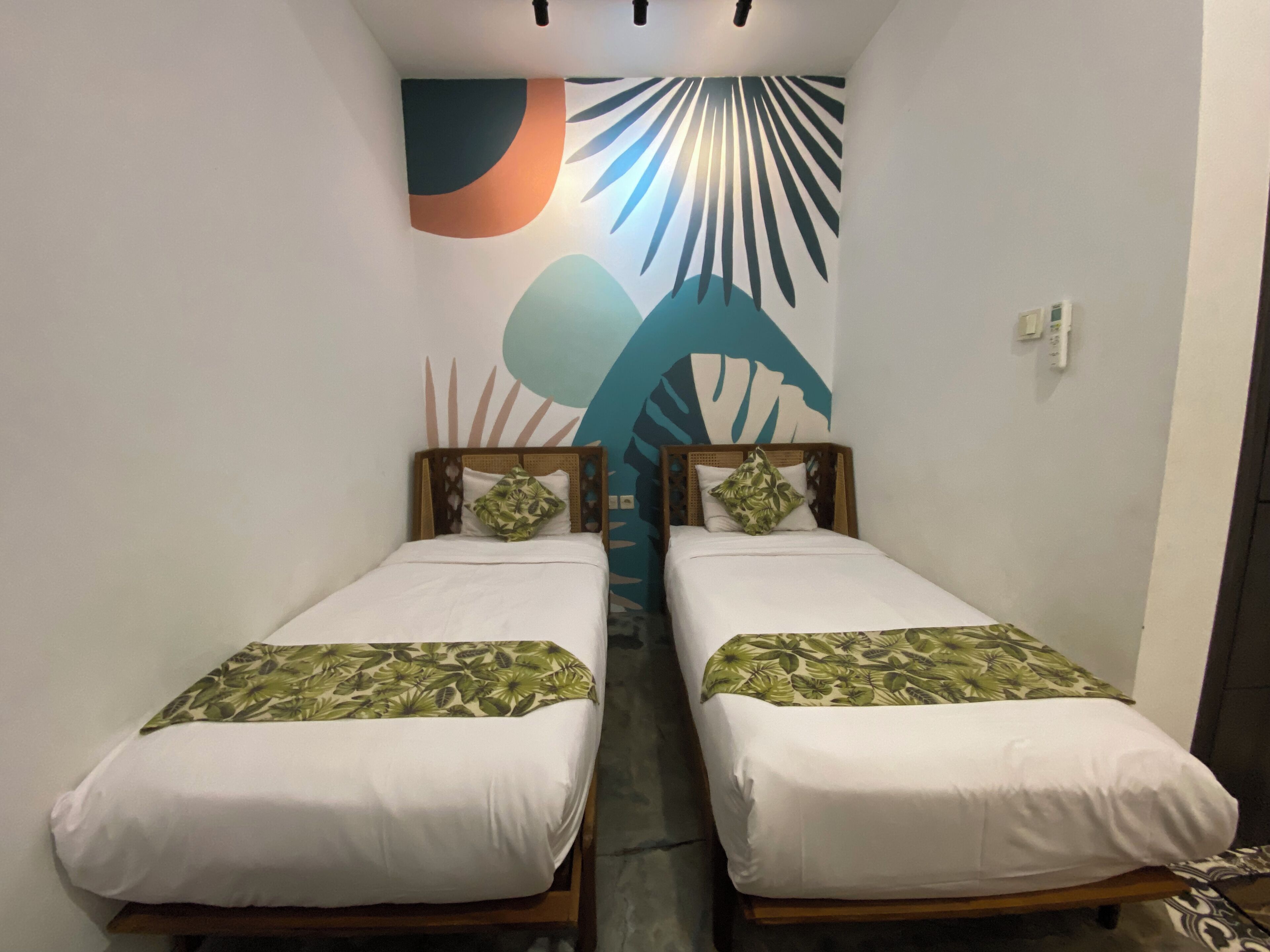 Een kamer met twee eenpersoonsbedden, elk met een witte matras en een bedspreide met bladpatroon. De muren zijn versierd met een groot tropisch bladmuurwerk.