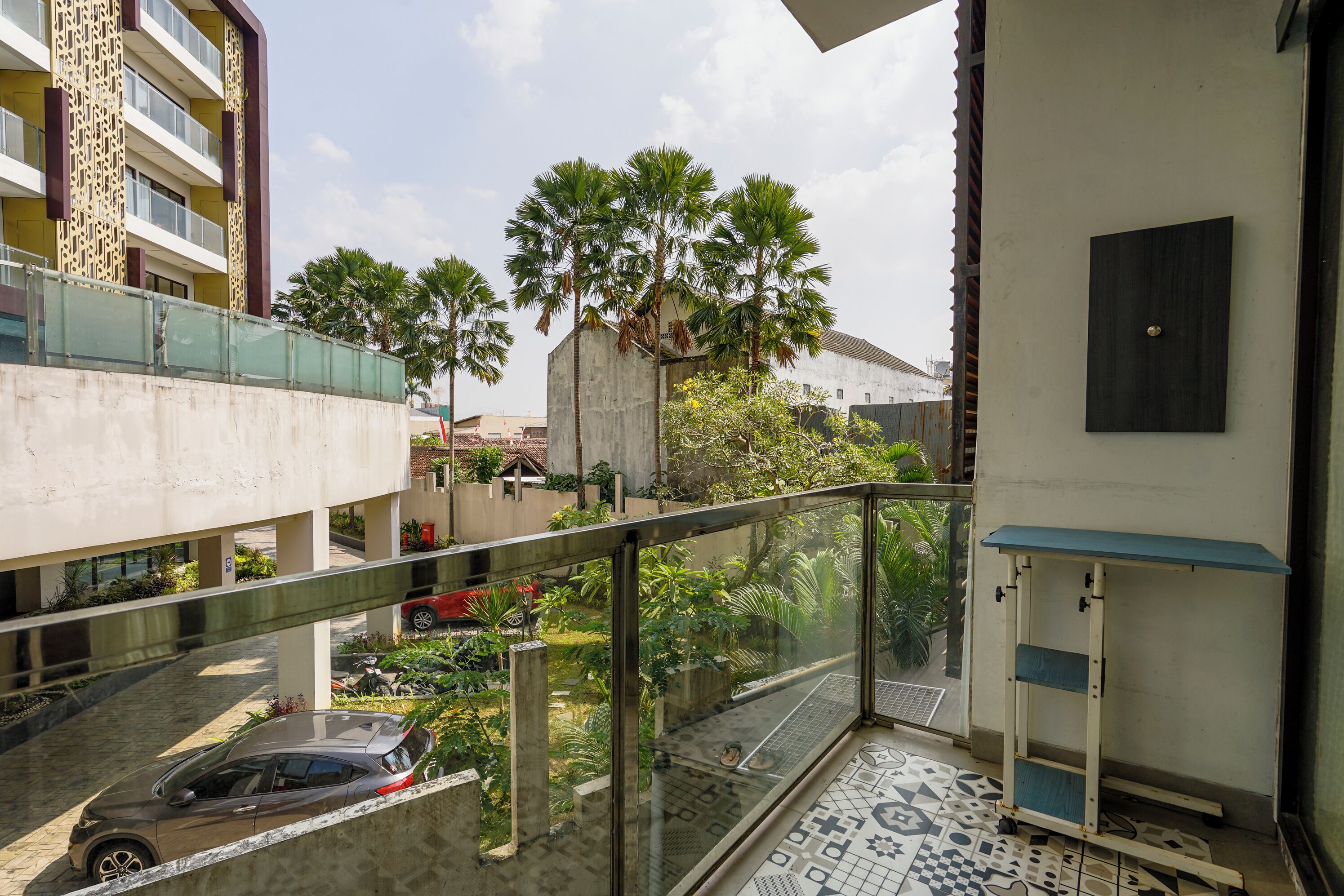 Un balcón residencial con barandilla de vidrio, piso de baldosas y vista a una calle con coches estacionados y un árbol.