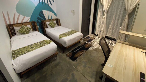 A room with two single beds, a wooden desk, a chair, a wall with a leaf pattern, and a door leading to another room.