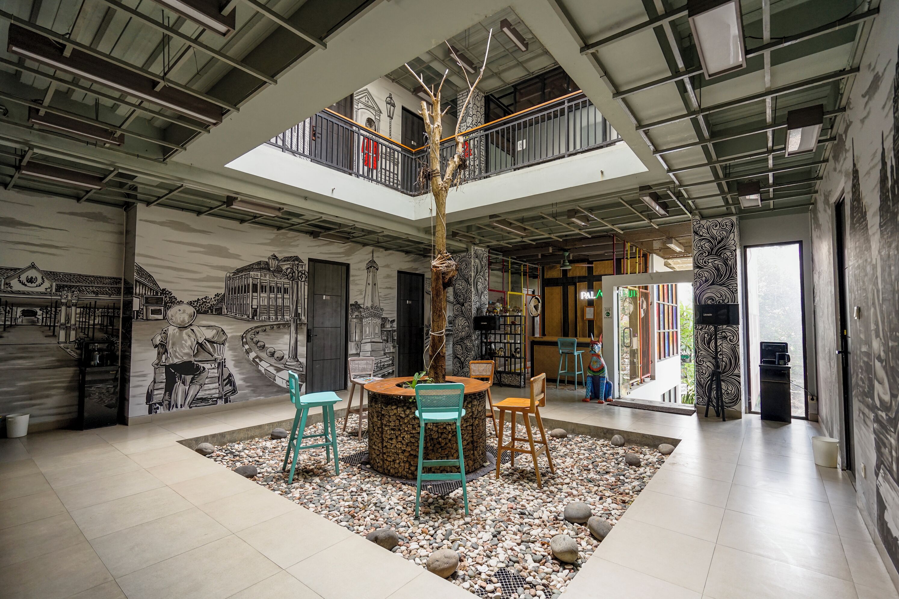 Un interior moderno con un árbol central, zona de estar y una escalera que lleva a un balcón.
