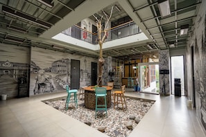 Un interior moderno con un árbol central, zona de estar y una escalera que lleva a un balcón.