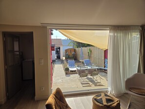 A room with a sliding glass door leading to an outdoor patio area with a table and chairs, a small garden, and a red building in the background.
