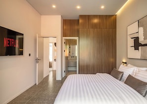 A modern bedroom with a large bed, a wooden accent wall, a mounted TV, and a hallway leading to a bathroom. - Elegant 2BR Private Pool Villa Umalas (Badung)