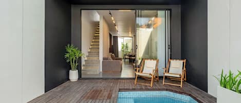 A modern outdoor pool area with a wooden deck, two lounge chairs, and a staircase leading to an interior space.