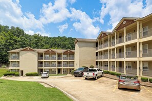 A multi-story hotel with balconies, a parking lot, and a forested hill in the background. - Lake Martin Condo Getaway with Pool! (Alexander City)