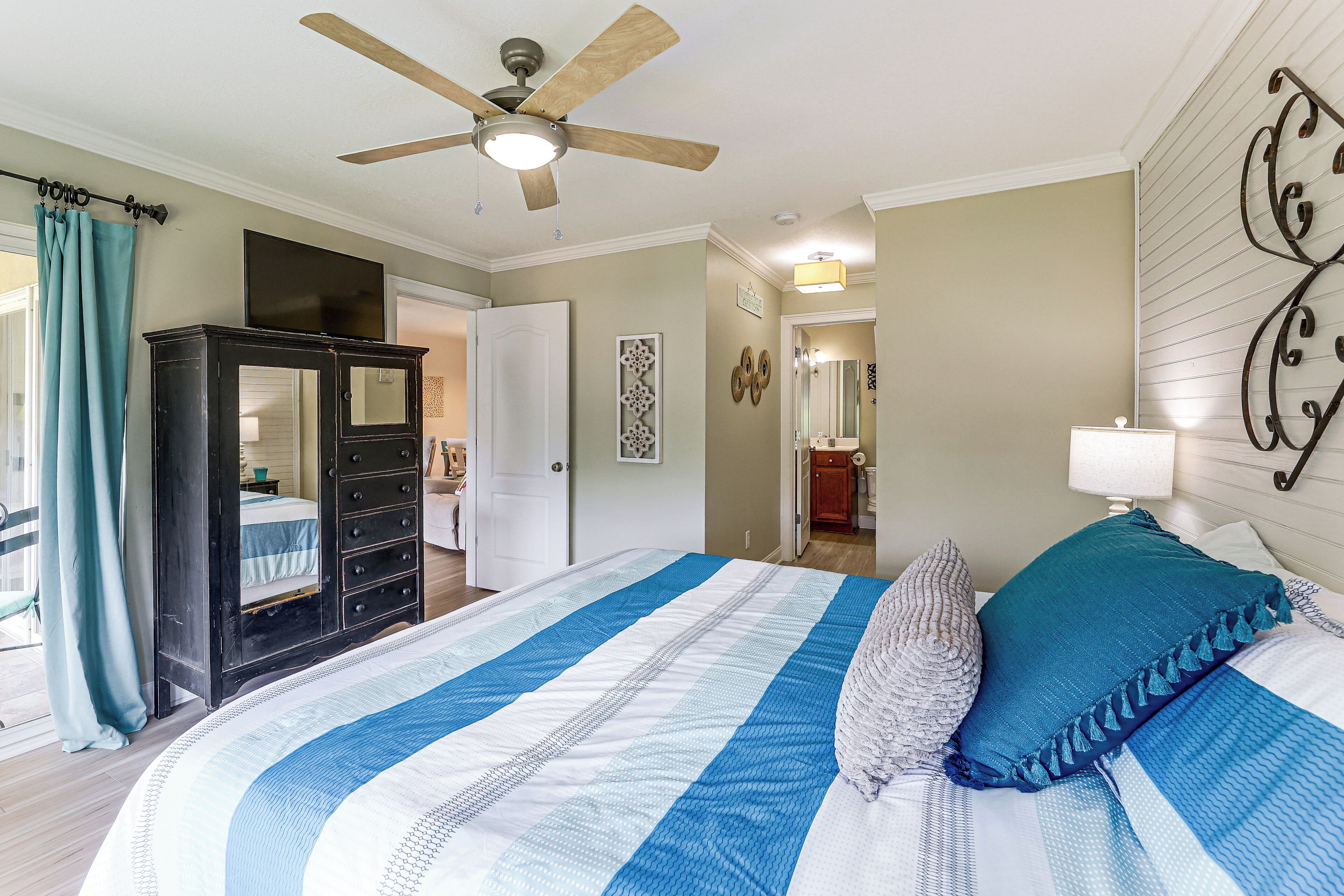 Une chambre à coucher avec un lit, un ventilateur de plafond, une armoire et une salle de bain visible à travers une porte ouverte.