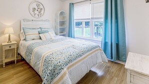 Un dormitorio con cama, mesita de noche, lámpara, decoración en la pared, ventana con cortinas y vistas al exterior.