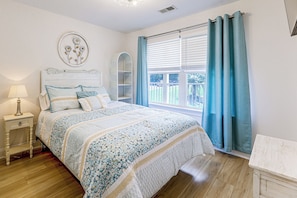 A bedroom with a bed, bedside table, lamp, decorative wall art, window with curtains, and a view of greenery outside. - Lake Martin Condo Getaway with Pool! (Alexander City)