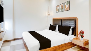A modern bedroom with a wooden bed frame, white bedding, a bedside table with a vase of flowers, and a wall-mounted light fixture. - Super Collection O Udaipur City (Udaipur)