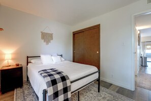 A bedroom with a bed, a nightstand, a lamp, a macrame wall hanging, and a wooden door. - Walk to Capitola Village: Condo w/ 2 Surfboards (Capitola)
