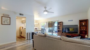 Ein Wohnzimmer mit einer Couch, einem Fernseher und einem Deckenventilator. Im Hintergrund ist eine Küchenzeile erkennbar.