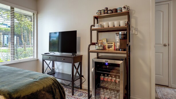Ein Zimmer mit Fernseher auf einem Schreibtisch, einem Regal mit Tassen und einer Kaffeemaschine sowie einem mit Getränken gefüllten Kühlschrank.
