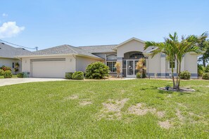 A residential house with a garage, a well-maintained lawn, and a variety of landscaping including shrubs and a palm tree. - Boat Dock & Outdoor Kitchen! Port Charlotte Escape (Port Charlotte)
