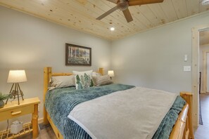 Un dormitorio con una cama, dos mesitas de noche, un ventilador de techo, un cuadro en la pared y una puerta a otra habitación.