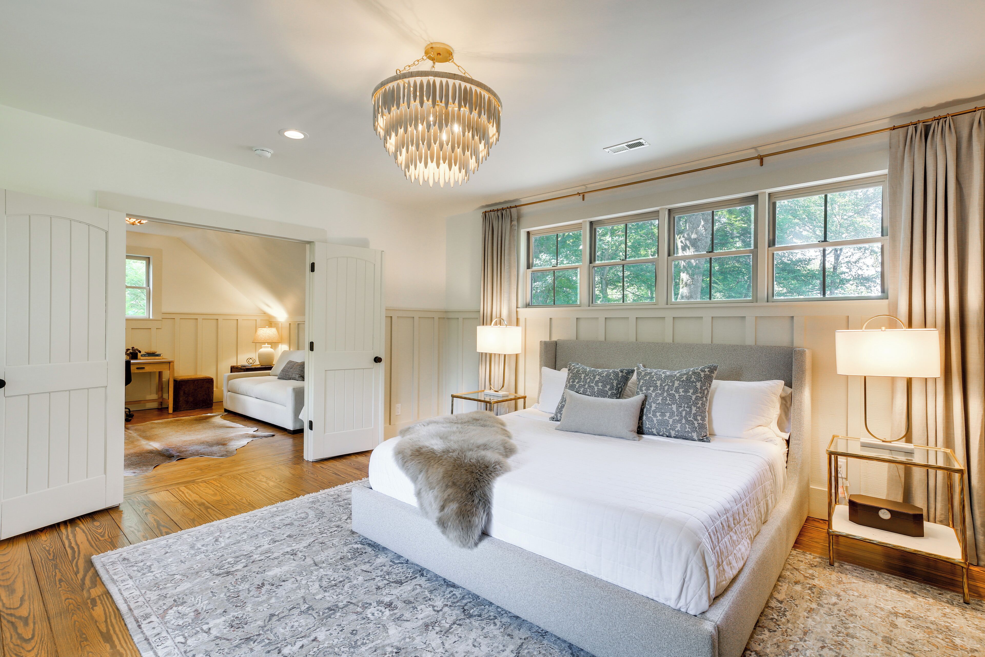 A spacious bedroom with a large bed, a chandelier, and a view of trees through the window.