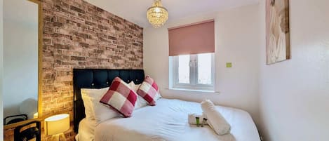 Une chambre d’hôtel avec un mur d’accent en briques, un lit avec une literie blanche et des oreillers à carreaux, une lampe de chevet et une fenêtre donnant sur un paysage agréable.