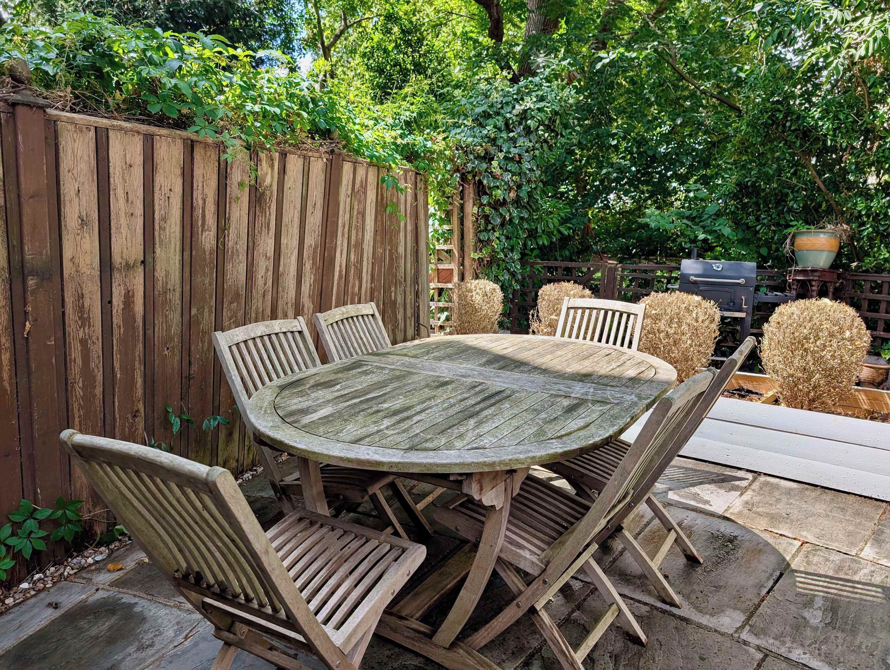 A wooden outdoor dining table with six chairs, surrounded by a wooden fence and lush greenery.