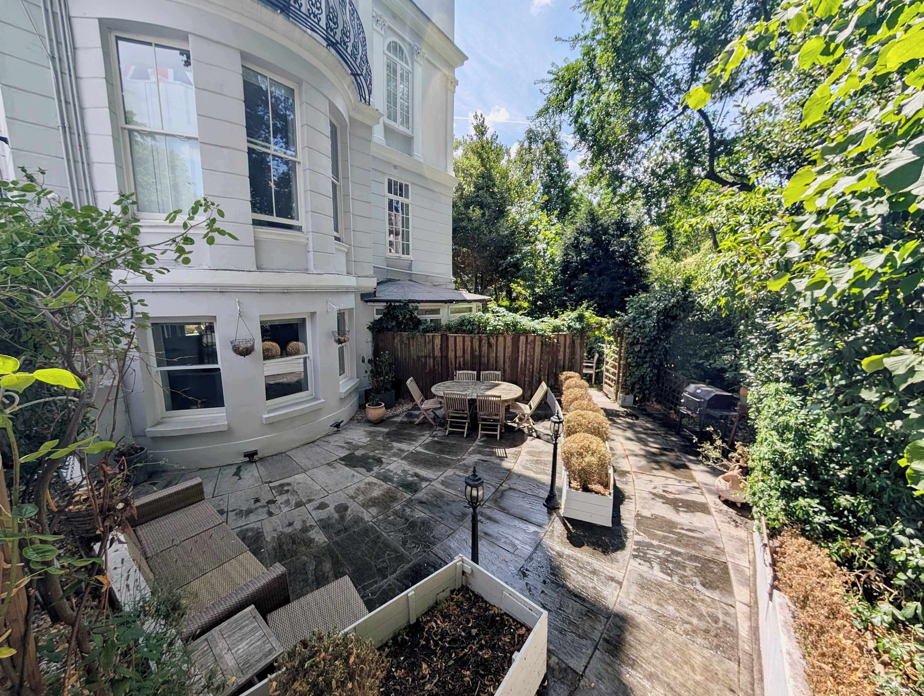 Eine Terrasse mit Holztisch und -stühlen, umgeben von einem weiß gestrichenen Gebäude mit großen Fenstern und einem Holzzaun.