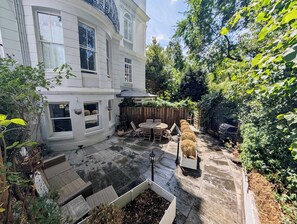 Eine Terrasse mit Holztisch und -stühlen, umgeben von einem weiß gestrichenen Gebäude mit großen Fenstern und einem Holzzaun.