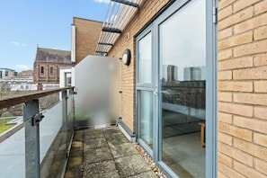 Uma varanda com porta de vidro, guarda-corpo metálico e vista para um edifício ao fundo.