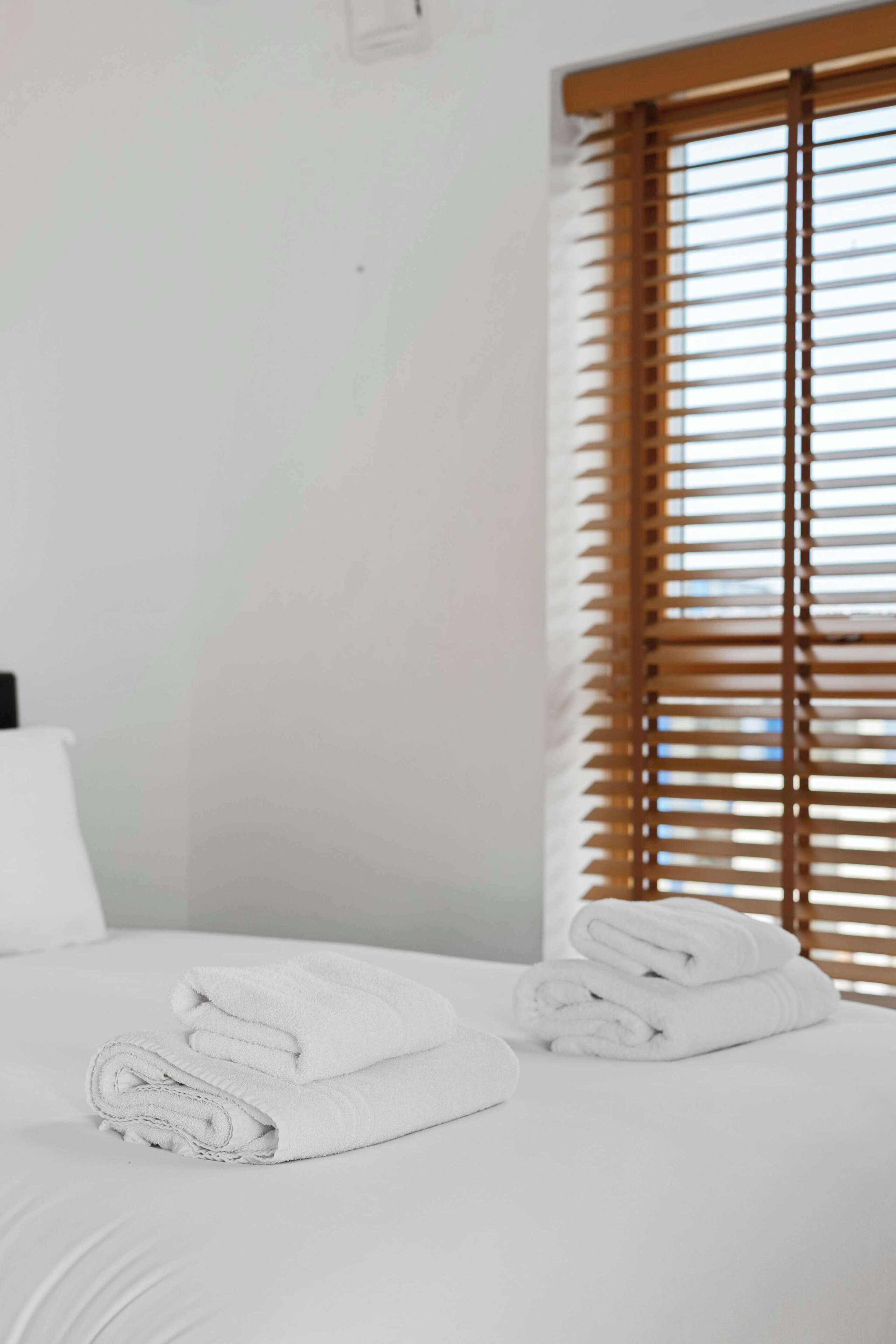 A hotel room with a bed, a window with blinds, and a folded towel on the bed.
