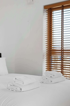 Quarto de hotel com cama, janela com persianas e uma toalha dobrada sobre a cama.