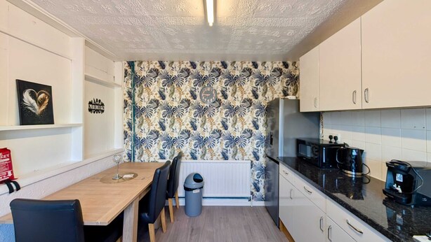 A compact kitchen with a wooden table, chairs, a black countertop, and a patterned wall. - 3bed Tilbury house with free parking (Tilbury)