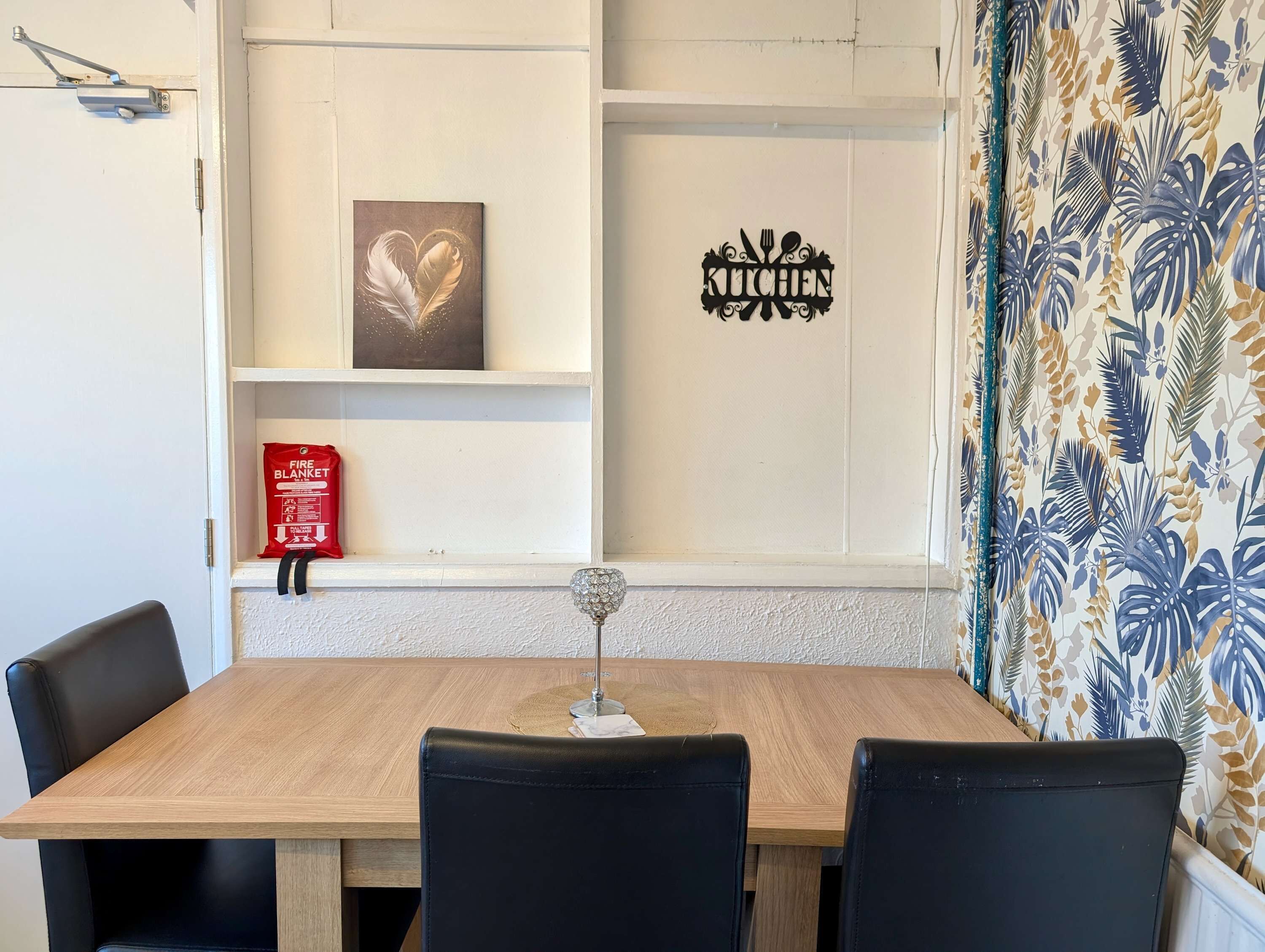 A dining area with a wooden table, black chairs, a fire blanket, a heart-shaped artwork, and a kitchen sign.