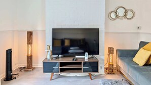 A modern living room with a flat-screen TV mounted on a white brick wall, a wooden entertainment unit, a grey sofa with yellow cushions, and a decorative wall piece. - 3bed Tilbury house with free parking (Tilbury)