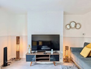 A modern living room with a flat-screen TV mounted on a white brick wall, a wooden entertainment unit, a grey sofa with yellow cushions, and a decorative wall piece. - 3bed Tilbury house with free parking (Tilbury)