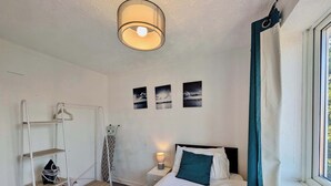 A bedroom with a bed, a bedside table with a lamp, a shelf with a bag, and a window with curtains. - 3bed Tilbury house with free parking (Tilbury)