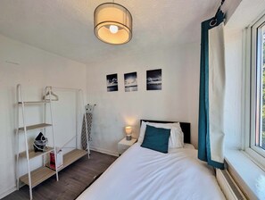 A bedroom with a bed, a bedside table with a lamp, a shelf with a bag, and a window with curtains. - 3bed Tilbury house with free parking (Tilbury)