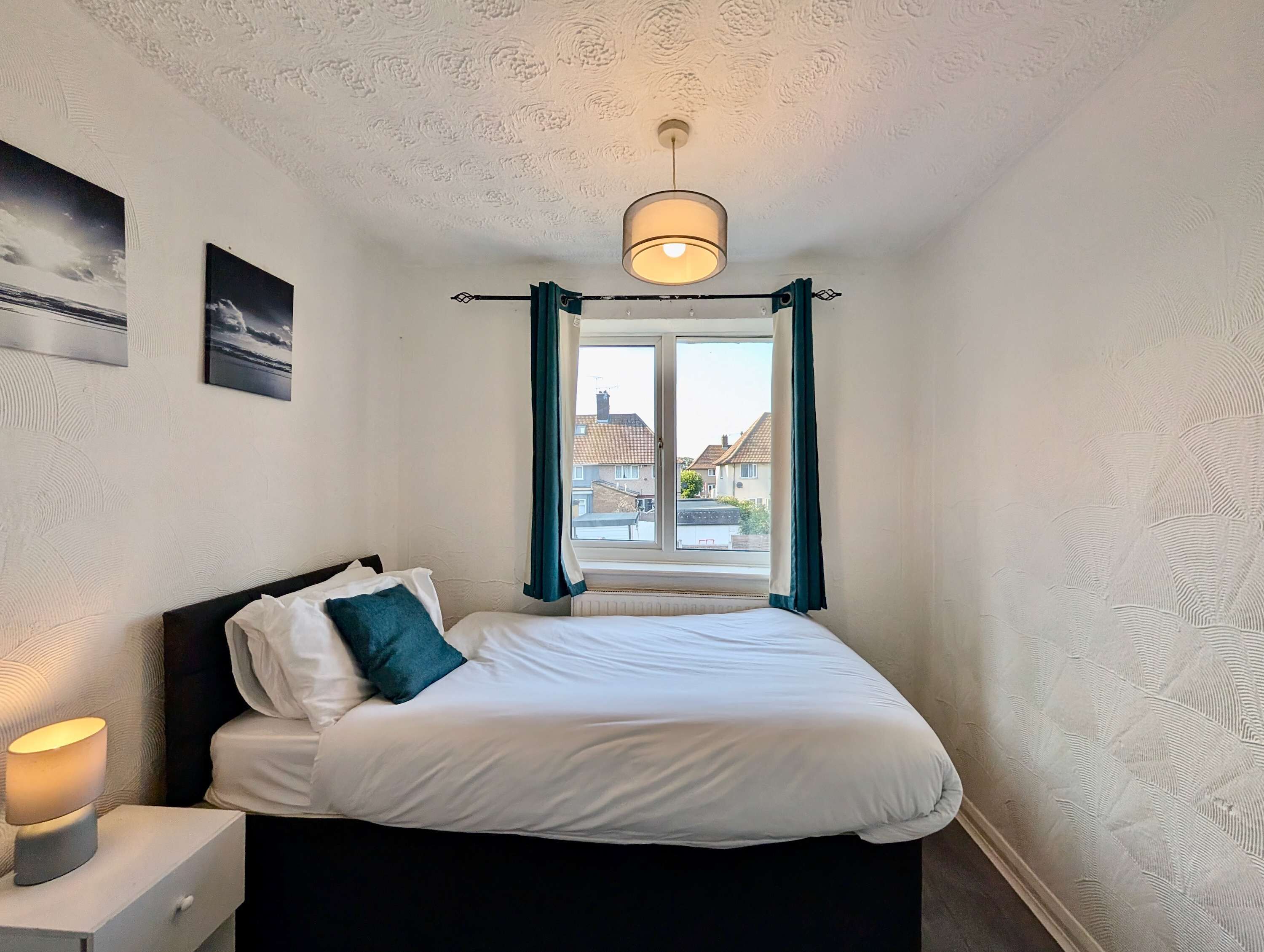 Un dormitorio con una cama, una mesita de noche, una lámpara, una ventana con cortinas y vistas a casas.