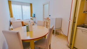 Hotellihuone, jossa on pyöreä ruokapöytä, tuolit, valkoisella päiväpeitteellä varustettu sänky, työpöytä tietokoneella, pieni keittokomero ja ikkuna, jossa on verhot.