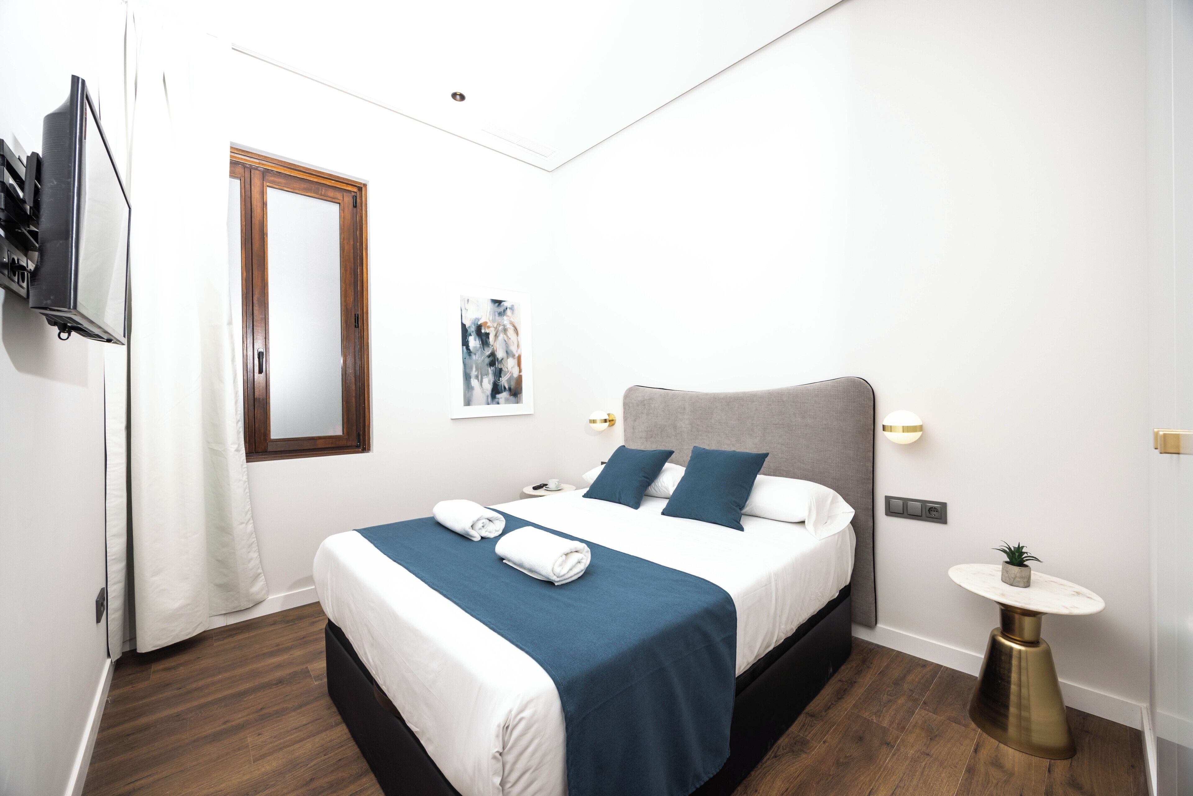 A hotel room with a bed, a bedside table, a small round table, a wall-mounted TV, and a window with curtains.