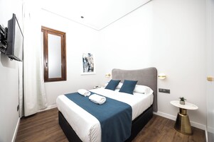 Habitación de hotel con cama, mesita de noche, una pequeña mesa redonda, televisor montado en la pared y ventana con cortinas.