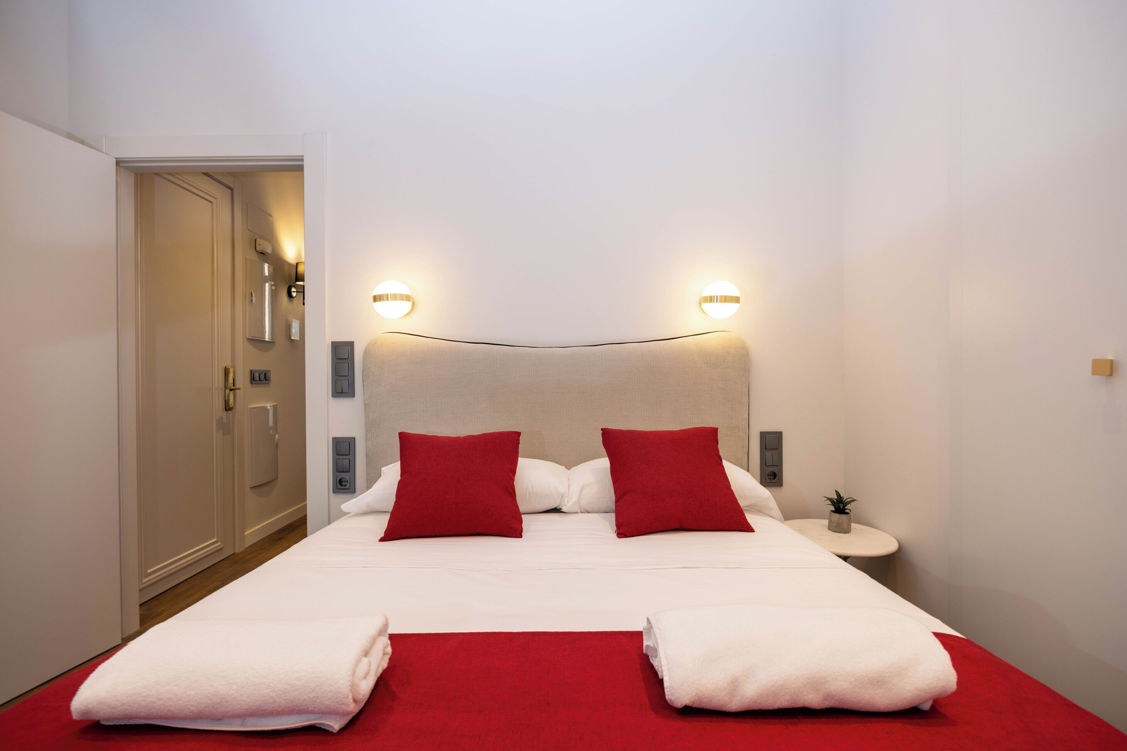 A hotel room with a bed, two red pillows, a white bedspread, two white towels, a small bedside table with a plant, and a door to the left.