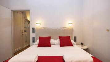 A hotel room with a bed, two red pillows, a white bedspread, two white towels, a small bedside table with a plant, and a door to the left.