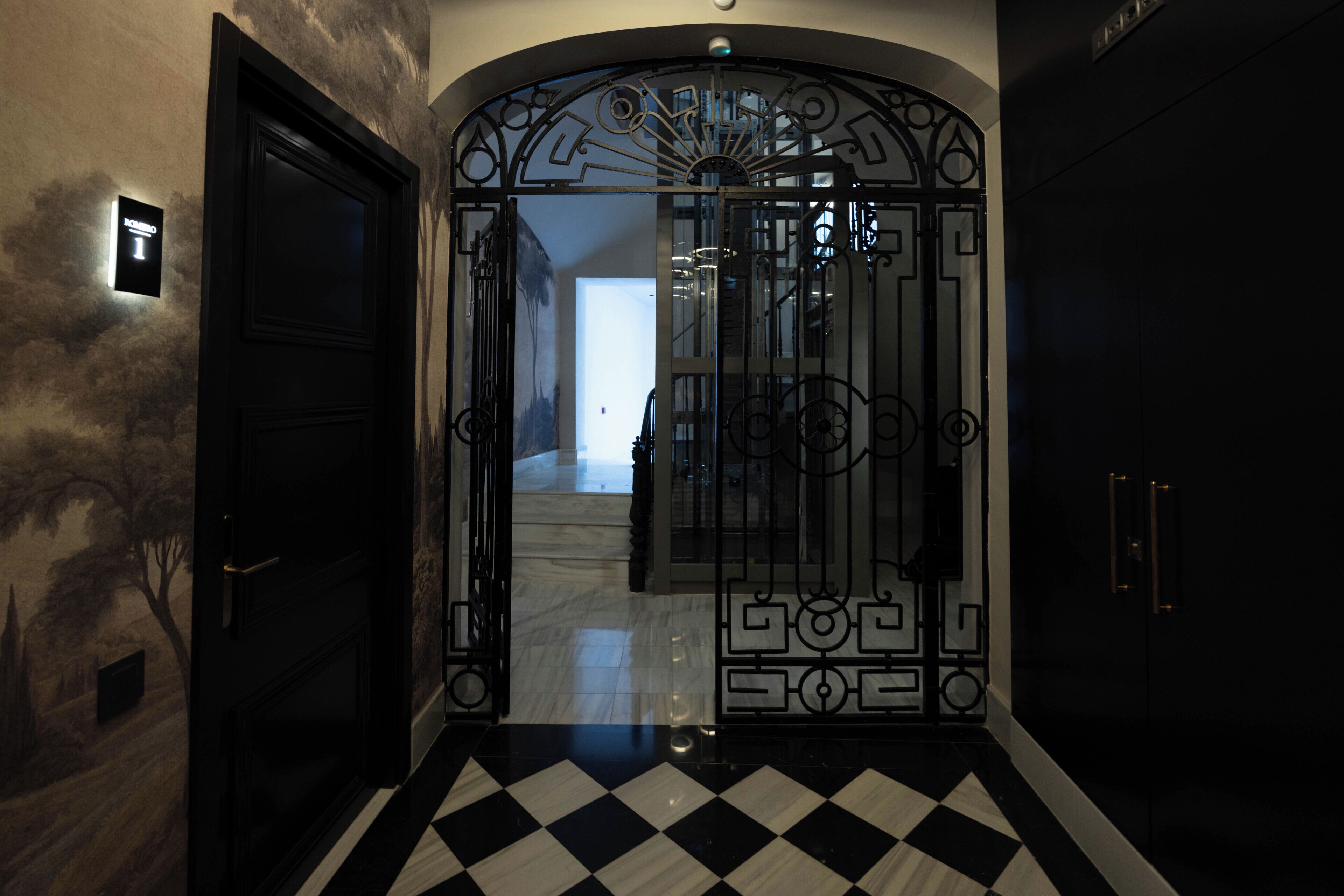 Un pavimento bianco e nero a scacchi che conduce a una stanza con una porta nera e un cancello metallico decorativo.