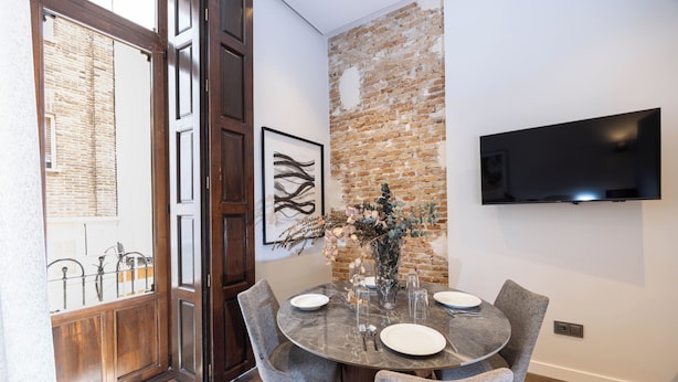 A dining area with a brick wall, a mounted TV, and a glass table with chairs.