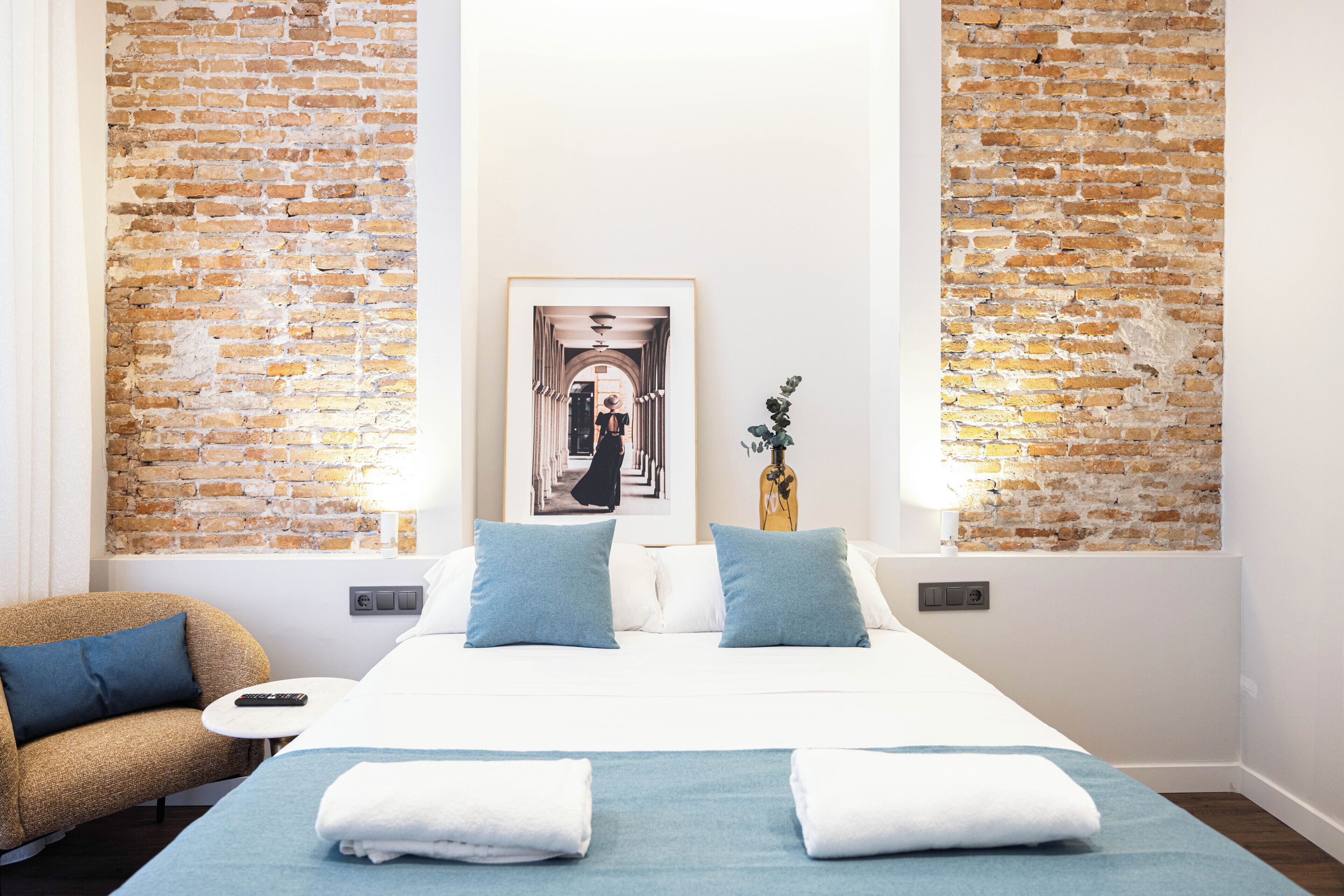A hotel room with a brick wall, a bed with blue bedding, a sofa, and a framed picture on the wall.