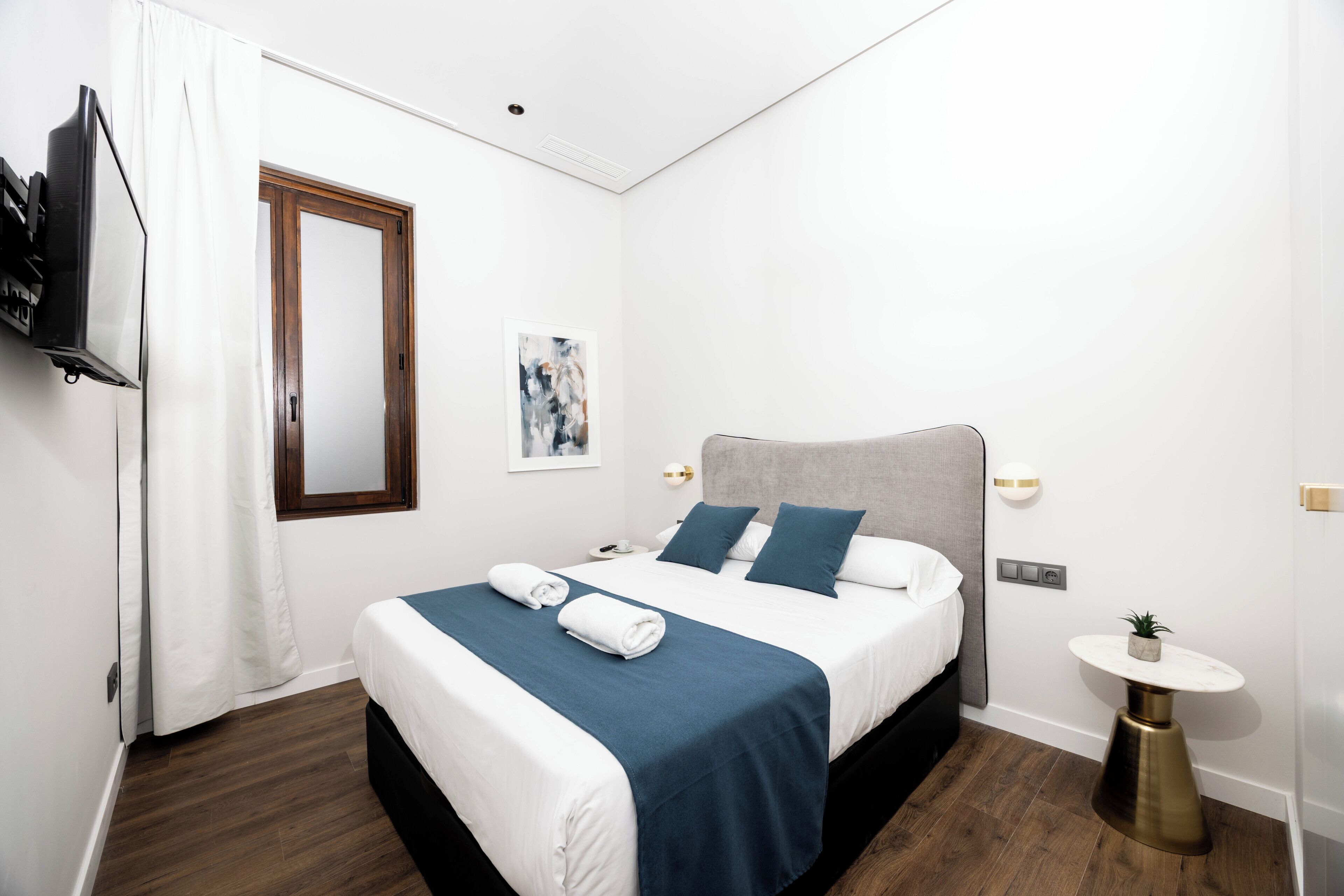 A hotel room with a bed, a bedside table, a small round table, a wall-mounted TV, and a window with curtains.