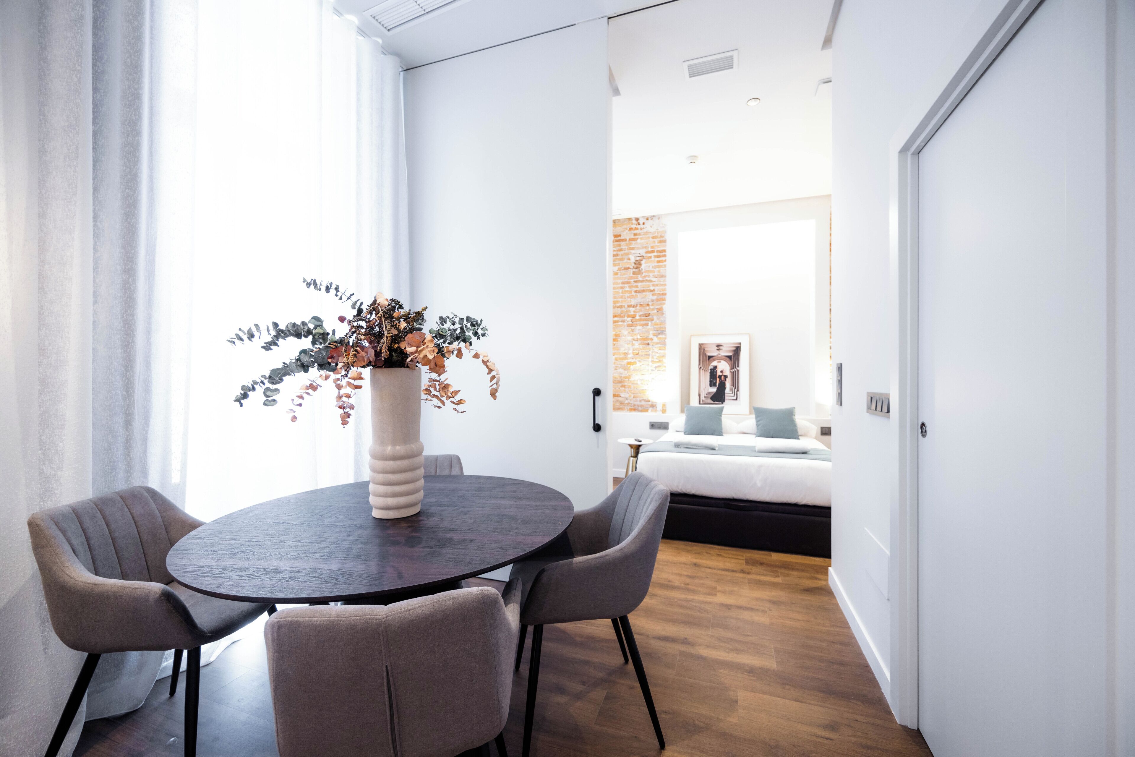A modern room with a round table, two chairs, a vase with dried flowers, and a bed with a blue pillow.
