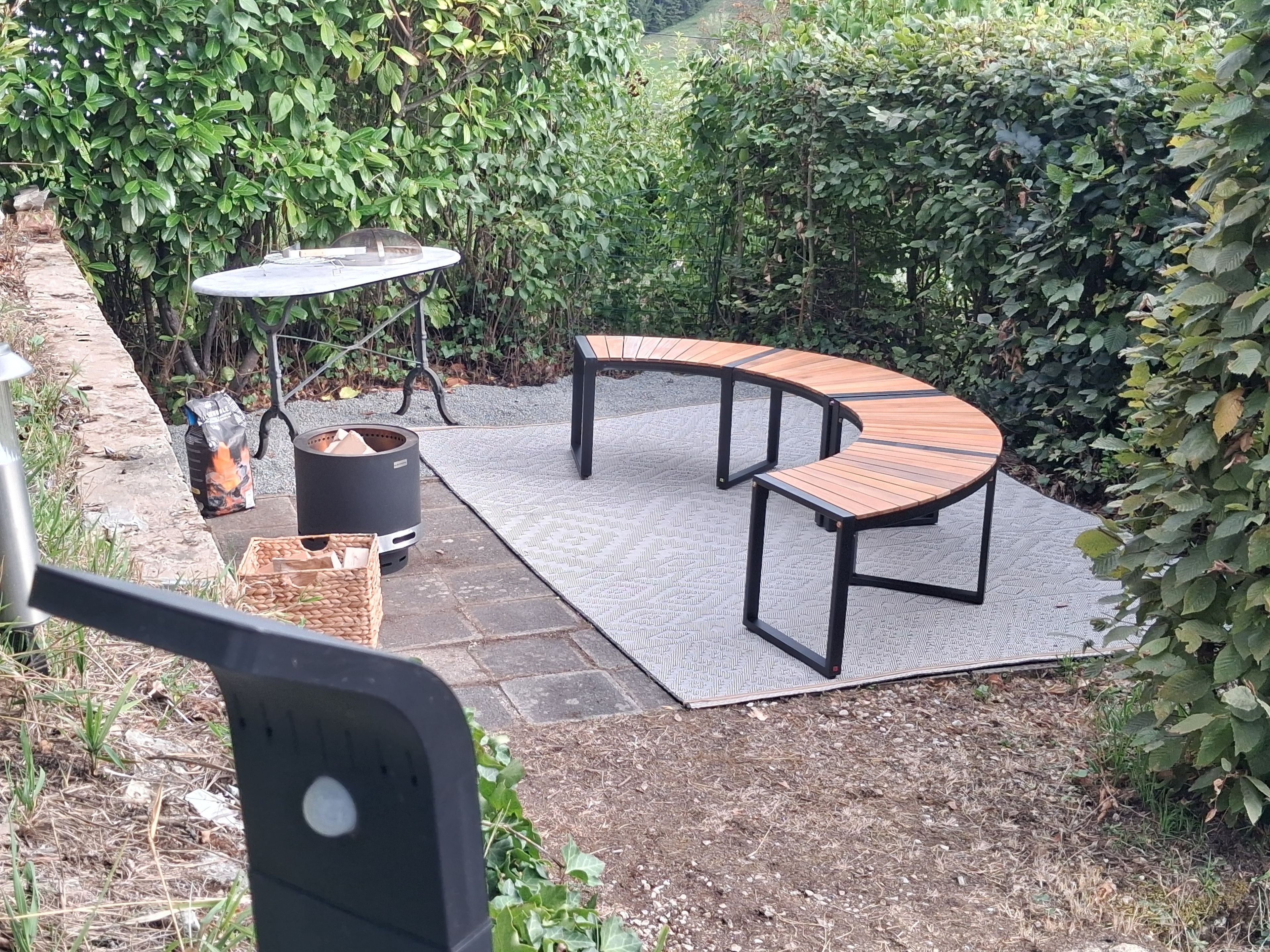 Une terrasse extérieure avec un banc en bois aux lignes courbes, une table ronde et un barbecue noir.