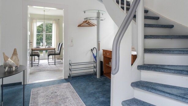 Een trap met een leuning, een eethoek met tafel en stoelen, en een uitzicht door een deur naar een balkon.