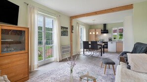 Een ruime woonkamer met een televisie, een eettafel met stoelen, een keuken op de achtergrond en grote ramen met uitzicht op het landschap.